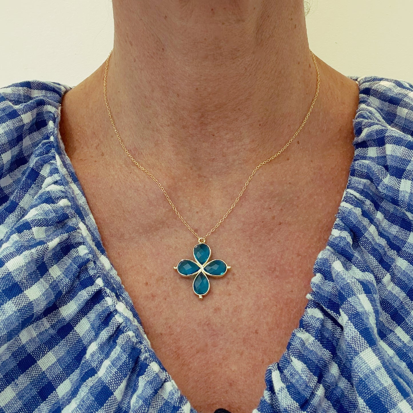 Necklace with a blue 4 petal flower shaped pendant worn by a person in a blue checkered shirt.