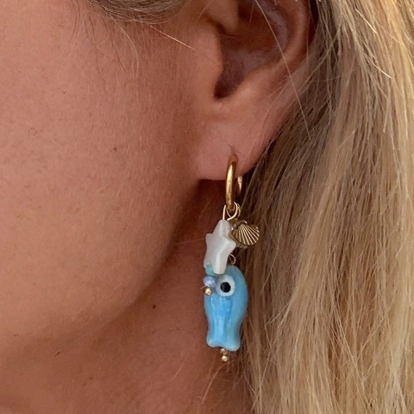 Close-up of an ear wearing a delicate gold hoop earrings with blue porcelain fish, mother of pearl white star and tiny gold shell charms