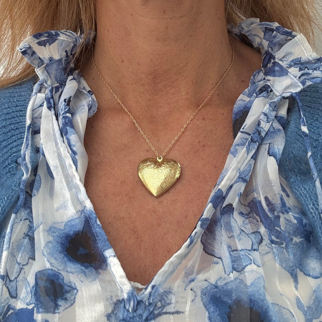 Gold heart-shaped locket necklace worn by a person in a blue and white floral top.