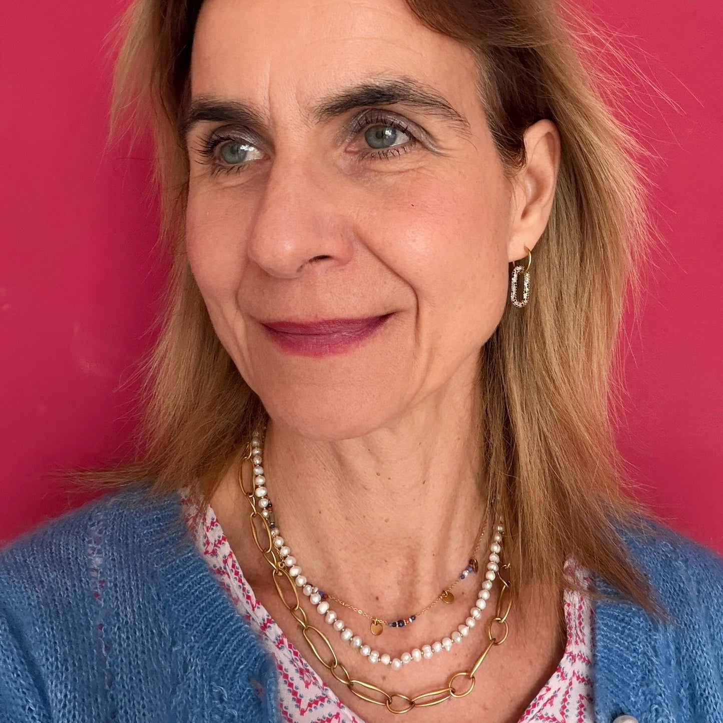 Woman with blonde hair and blue eyes wearing a blue cardigan and pearl necklace against a pink background