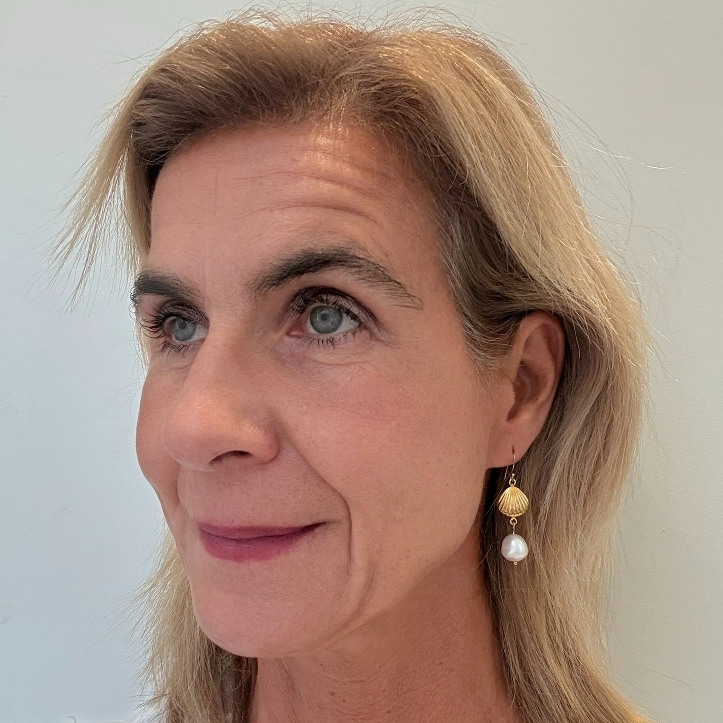 Woman with blonde hair wearing gold and pearl earrings against a plain background