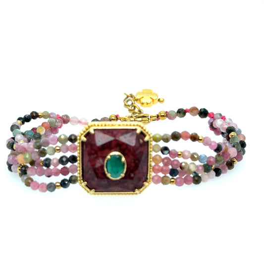 A multi-strand beaded bracelet featuring a large central faceted dark red gemstone with a gold border and a green oval inset, surrounded by multiple strands of small multicolored beads on a white background.