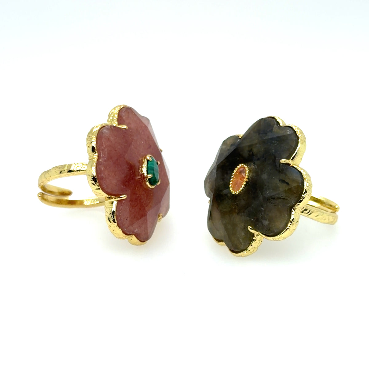 Two gold flower-shaped rings with cabochon gemstones, one in reddish-pink with a small green center and the other in dark brown with a small orange center, displayed on a white background.
