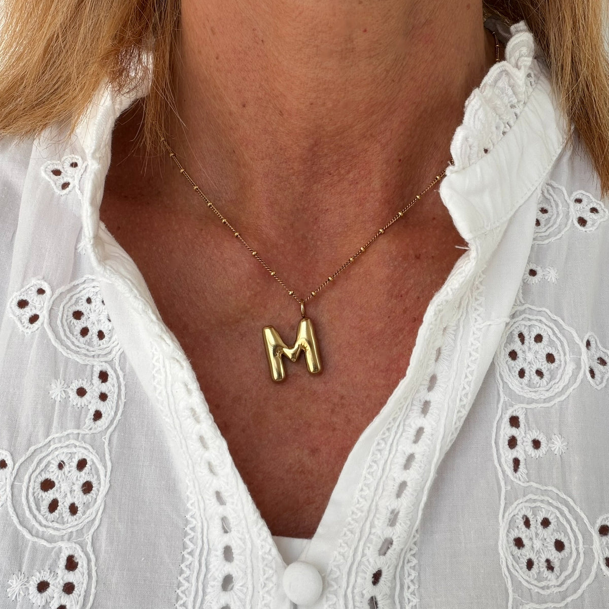 A close-up of a person wearing a delicate gold necklace with a polished letter 'M' pendant, styled with a white eyelet blouse.
