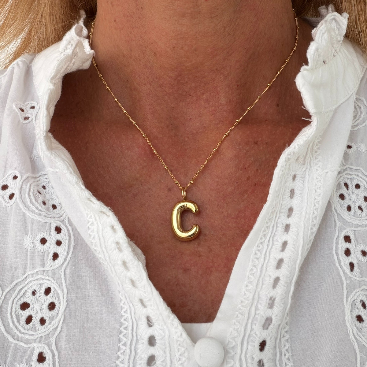 A close-up of a person's neck and chest wearing a delicate gold chain necklace with a polished lowercase letter 'c' pendant, paired with a white eyelet blouse.