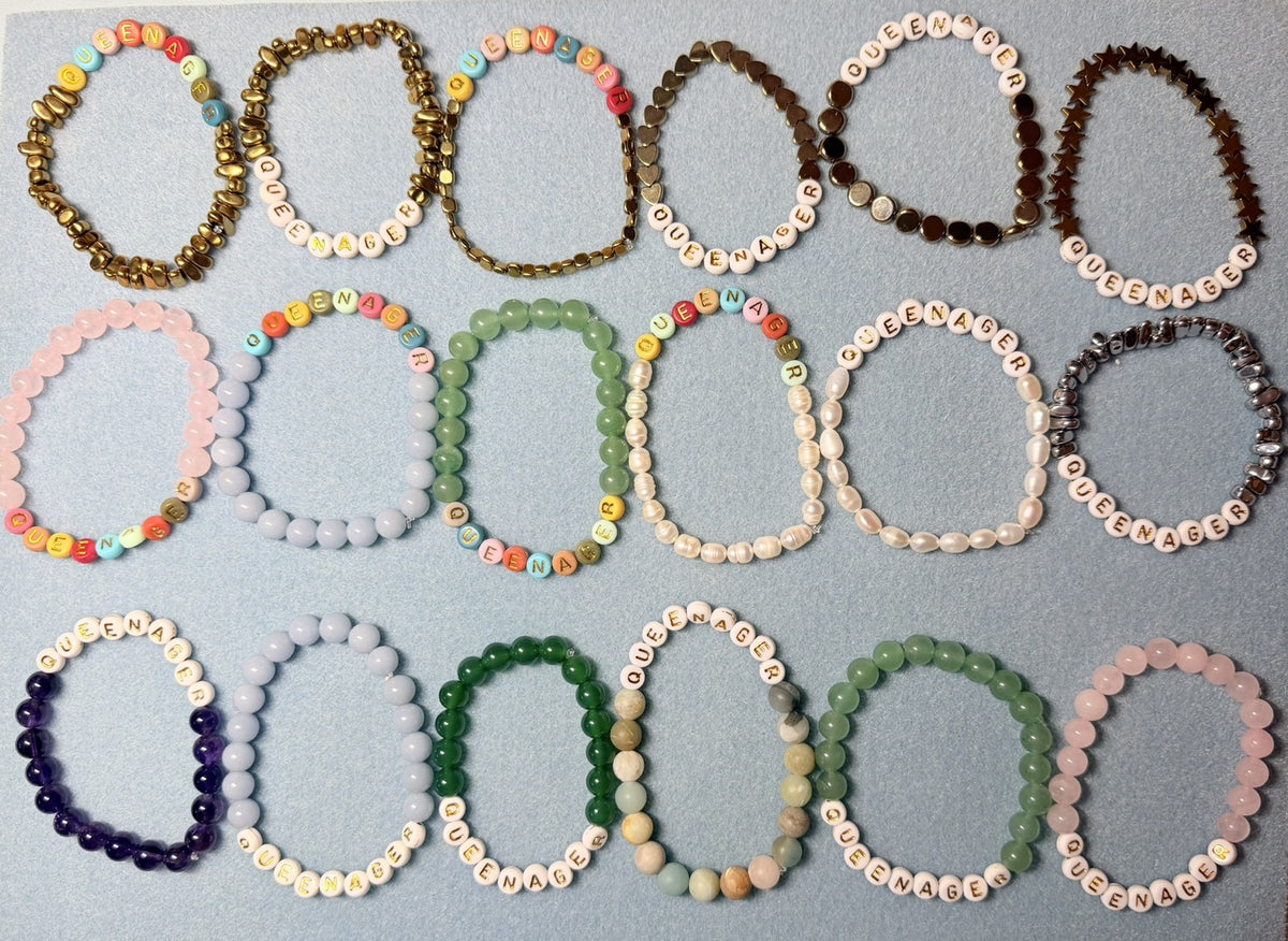 A collection of beaded stretch bracelets arranged in three columns on a light blue-gray background. Each bracelet features white round beads printed with colorful letters, paired with various colored stone beads in shades of pink, green, blue, purple, brown, and gold.